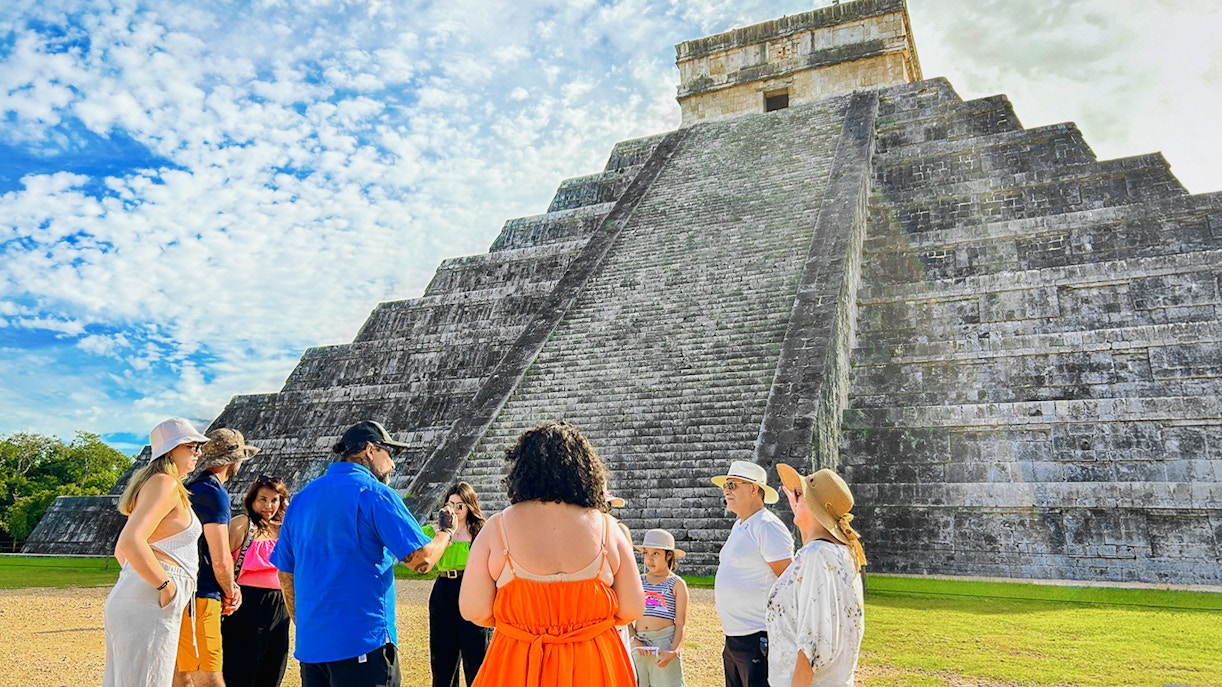 Tour group at Chichen Itza pyramid, Mexico, with guide explaining site history.