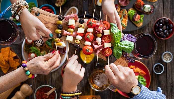 Hands sharing skewers of tapas with olives, cheese, and tomatoes at a table with wine.