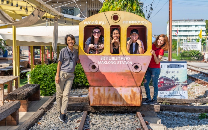 Visitors posing at Maeklong Railway Market, Thailand, near a train cutout.
