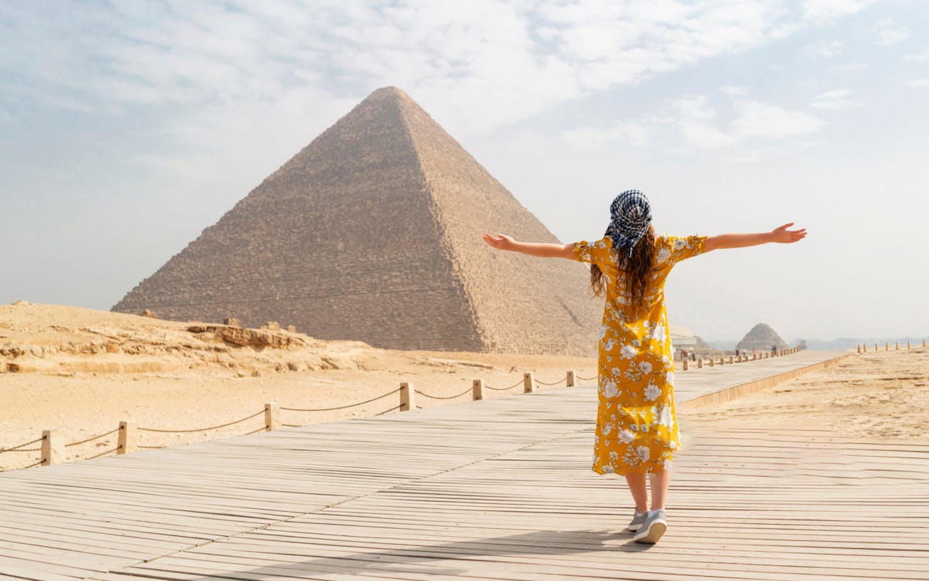 Person facing the Pyramid of Giza on a wooden walkway, arms outstretched.