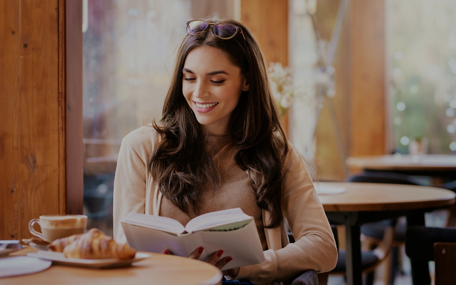 Woman reading a book in a cozy café with coffee and croissant on the table.