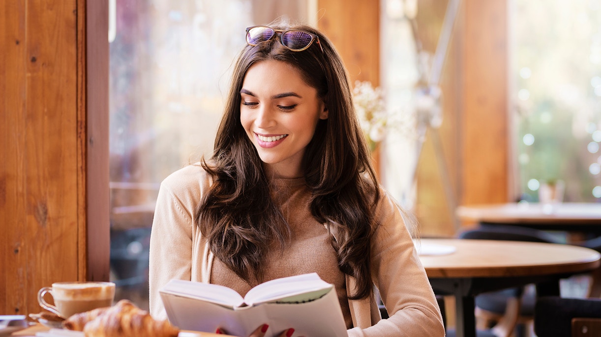 Woman reading a book in a cozy café with coffee and croissant on the table.