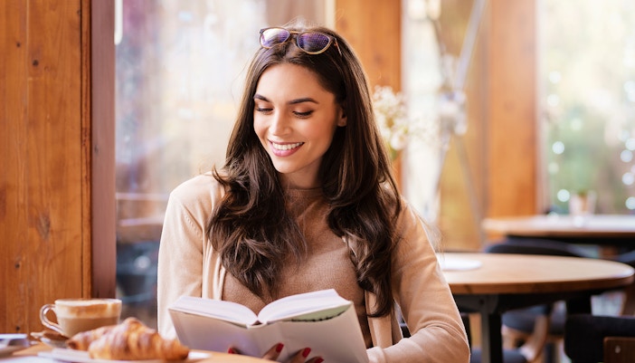 Woman reading a book in a cozy café with coffee and croissant on the table.