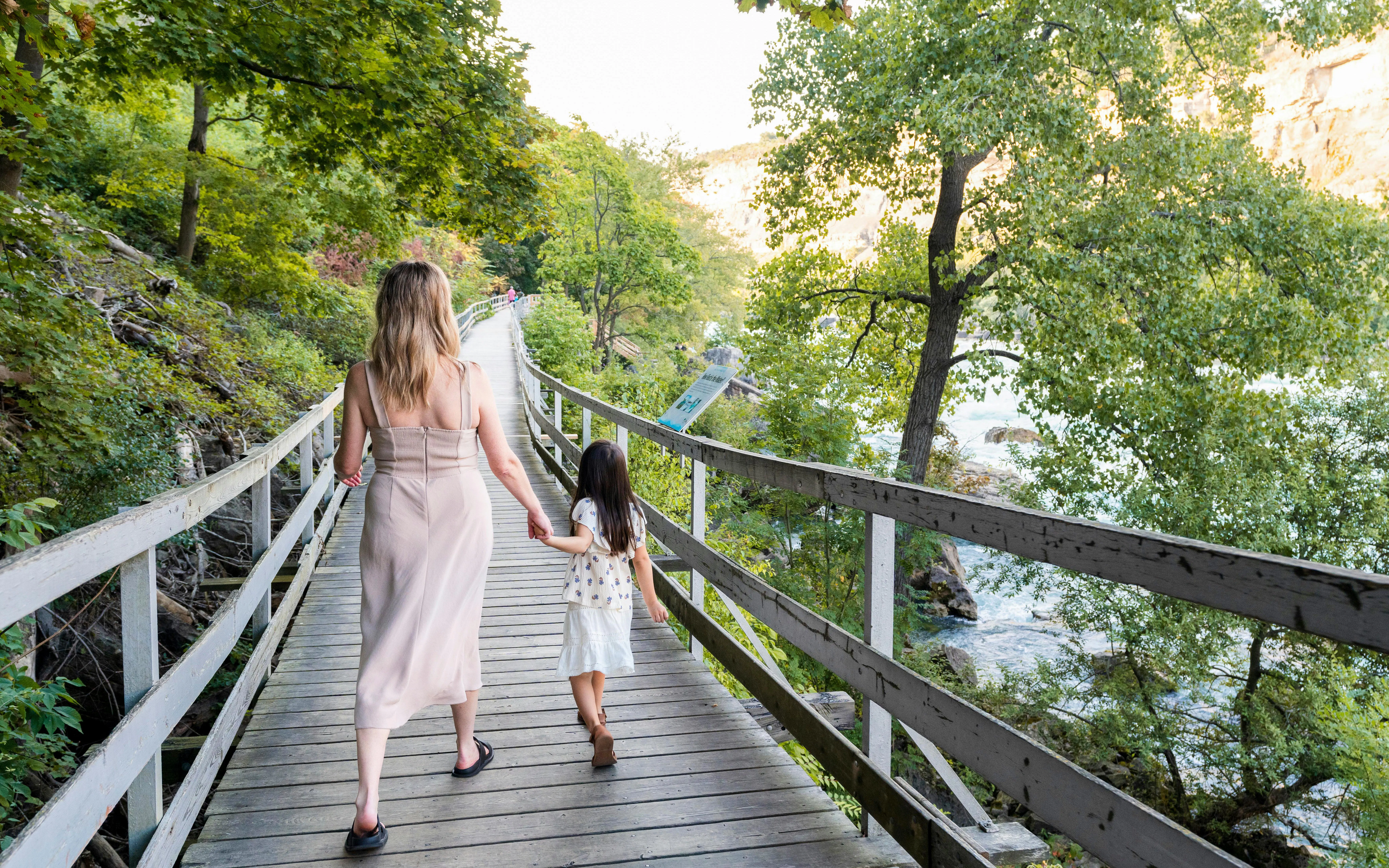 Visitors walking along a wooden path beside white water rapids in a forested area.