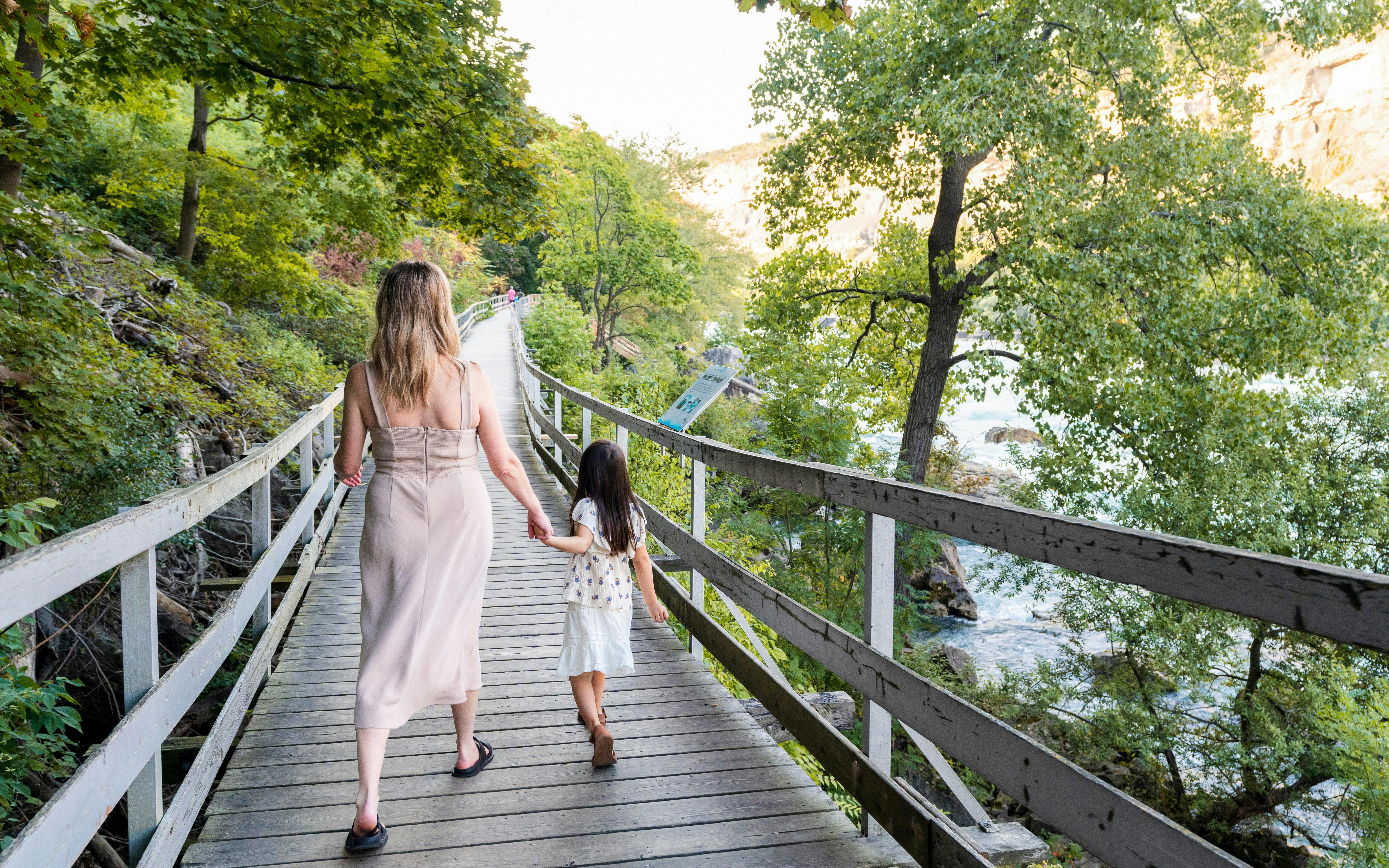Visitors walking along a wooden path beside white water rapids in a forested area.