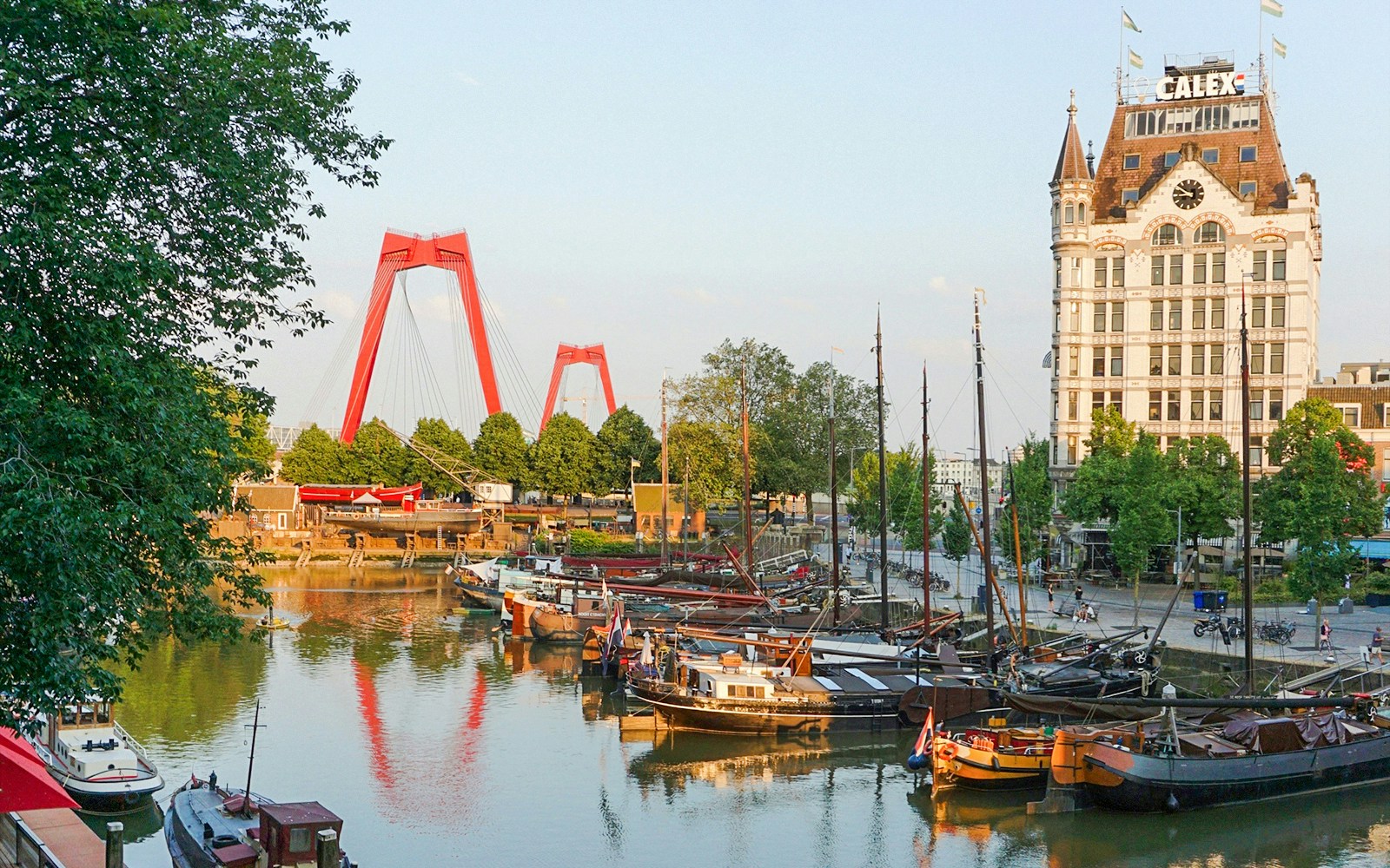 Historic boats docked at Wilton Haven, Rotterdam, with the iconic red bridge and White House building in view.