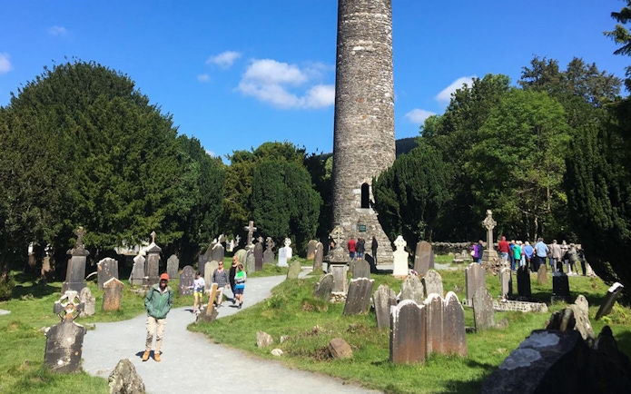 Glendalough round tower and cemetery with tourists exploring, part of Wicklow and Kilkenny tour from Dublin.