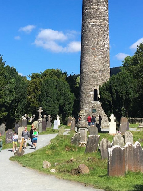 Glendalough round tower and cemetery with tourists exploring, part of Wicklow and Kilkenny tour from Dublin.