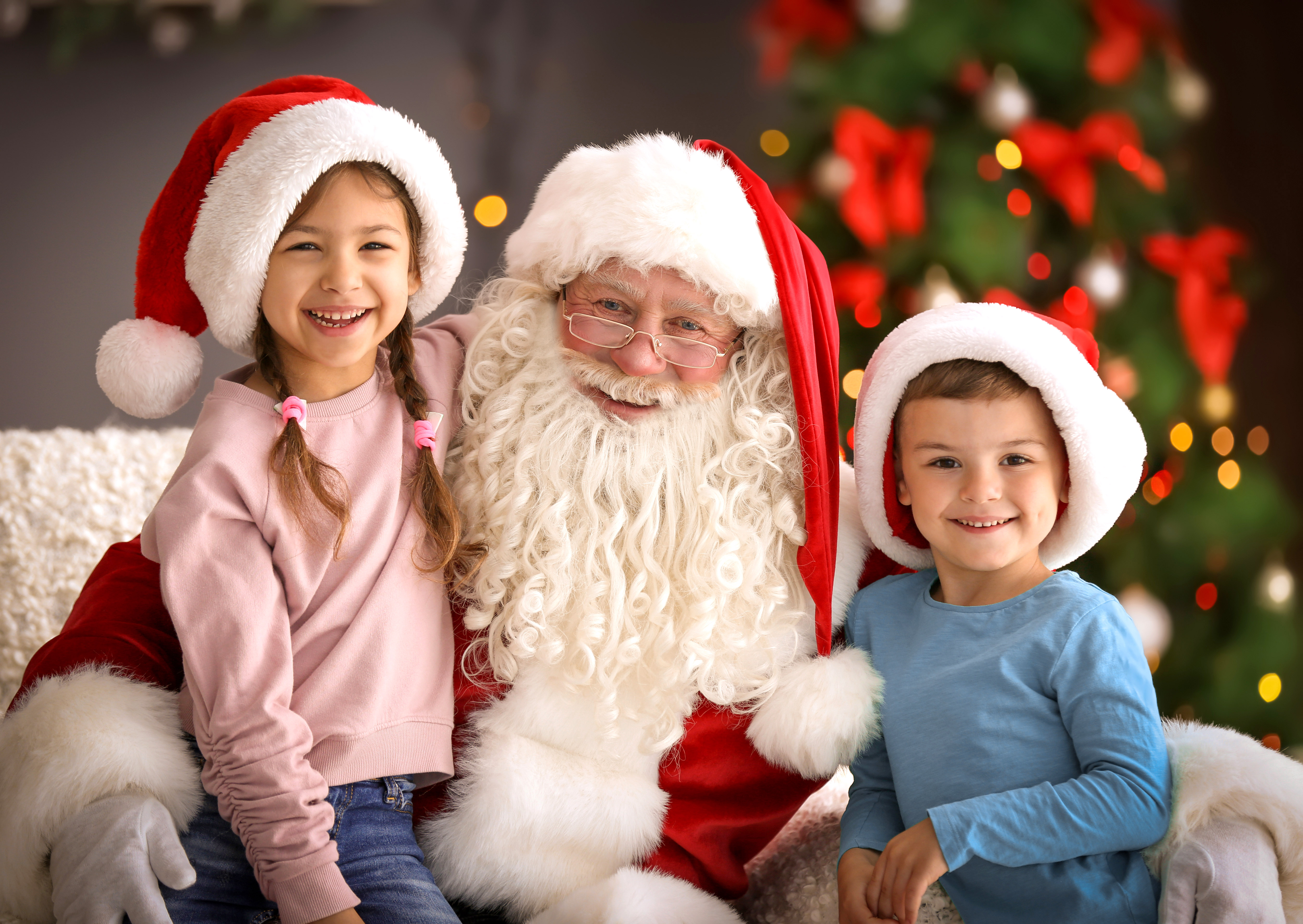 Santa Claus with two smiling children in festive hats by a decorated Christmas tree.