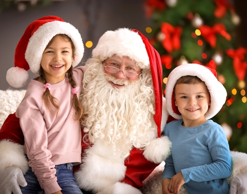 Santa Claus with two smiling children in festive hats by a decorated Christmas tree.