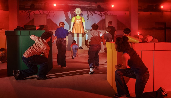 Participants in a Squid Game experience in London, hiding and moving towards a large doll.