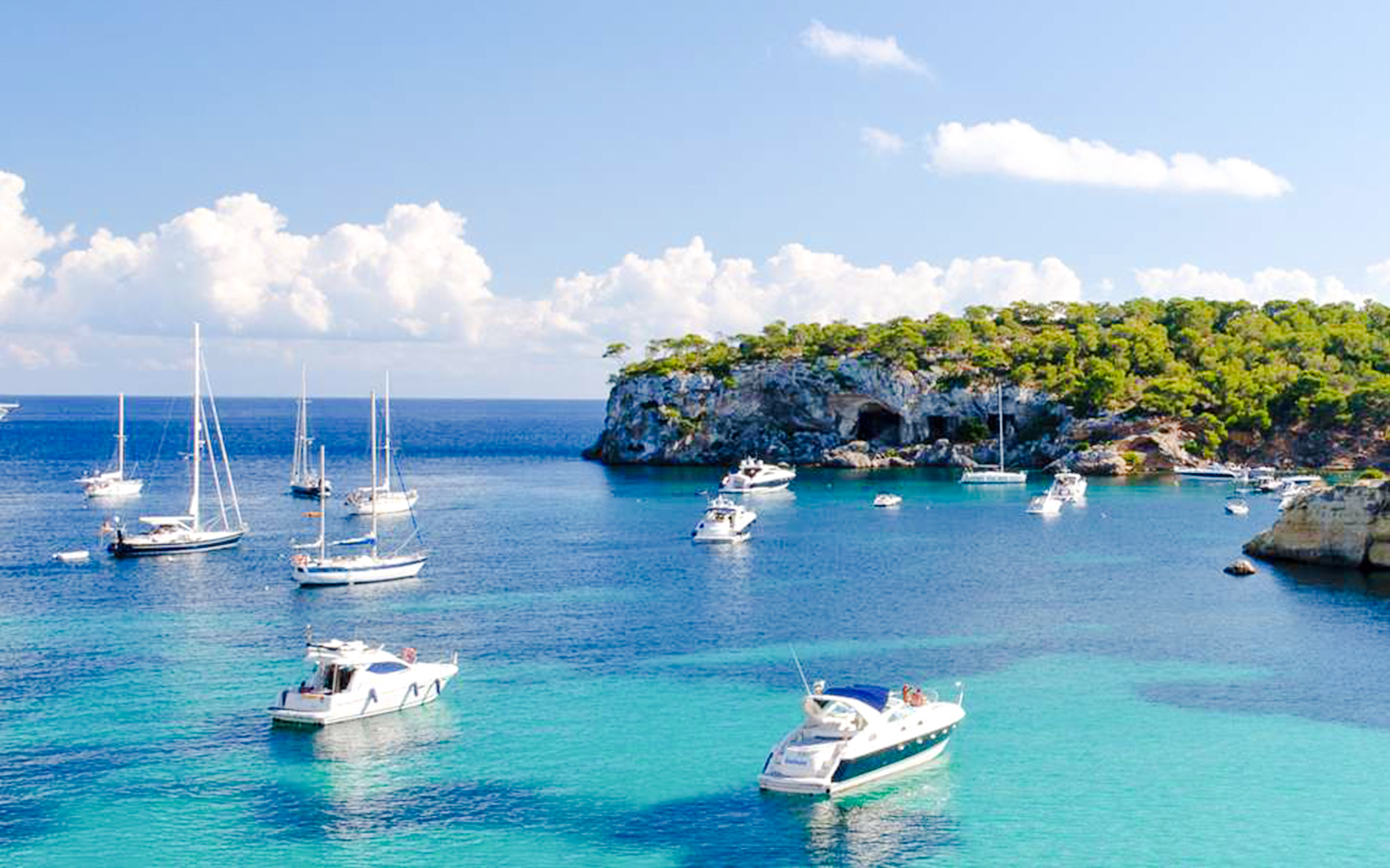 Sailboats and yachts anchored at Porto Cristo beach, Mallorca, with rocky cliffs and clear blue water.
