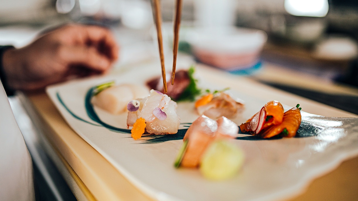 Sushi chef using chopsticks to prepare sashimi on a plate in a restaurant.
