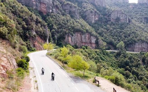 Motorcyclists riding BMW bikes on a scenic road in Montserrat, Spain.