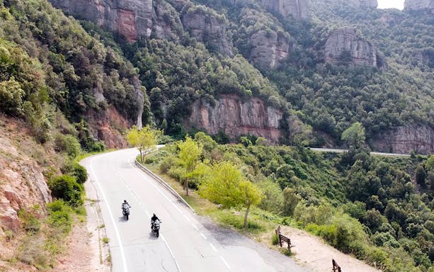 Motorcyclists riding BMW bikes on a scenic road in Montserrat, Spain.