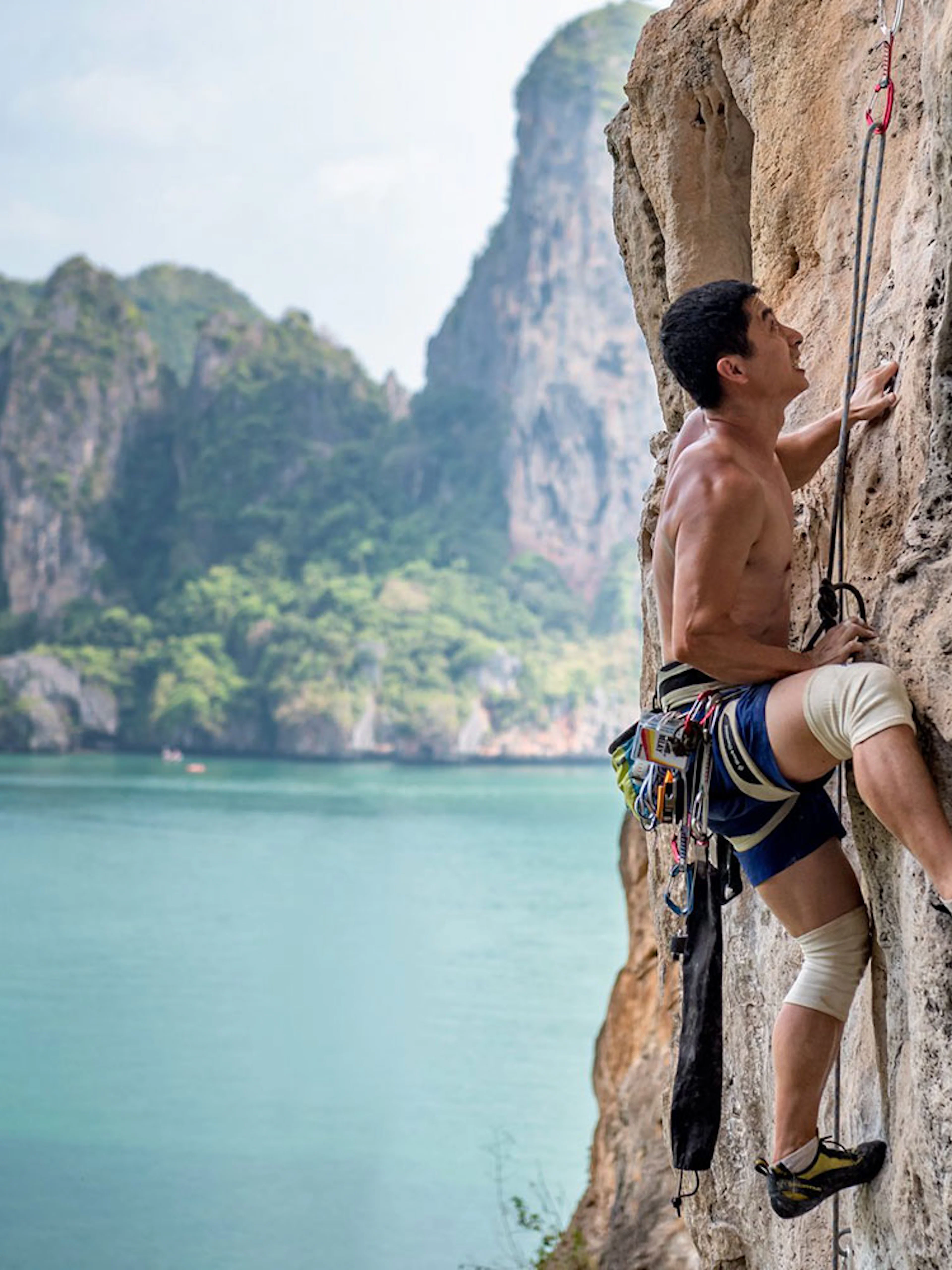 Rock climber scaling limestone cliff at Railay Beach, Krabi, with ocean and cliffs in background.