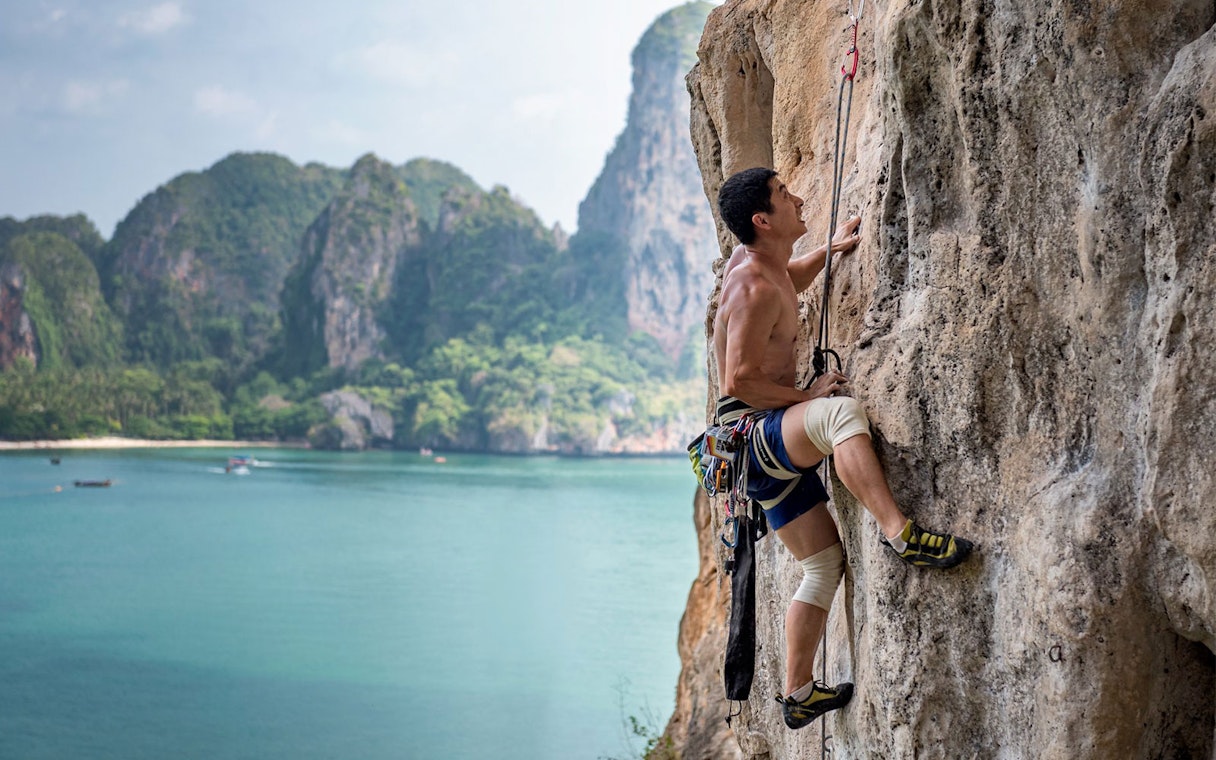 Rock climber scaling limestone cliff at Railay Beach, Krabi, with ocean and cliffs in background.