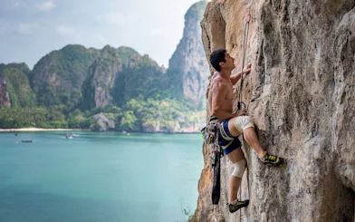 Rock climber scaling limestone cliff at Railay Beach, Krabi, with ocean and cliffs in background.