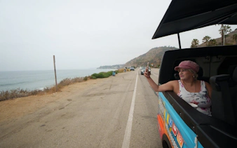 Open-air tour van on Pacific Coast Highway with passenger taking photo of Malibu coastline.