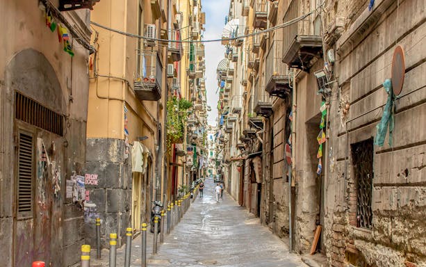 Narrow alley in Spaccanapoli, Naples, with colorful flags and rustic buildings.