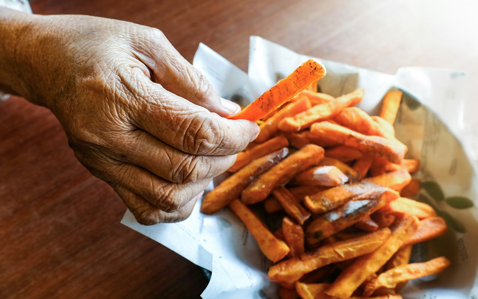 Elderly hand holding crispy sweet potato fry over a bowl of fried sweet potatoes.