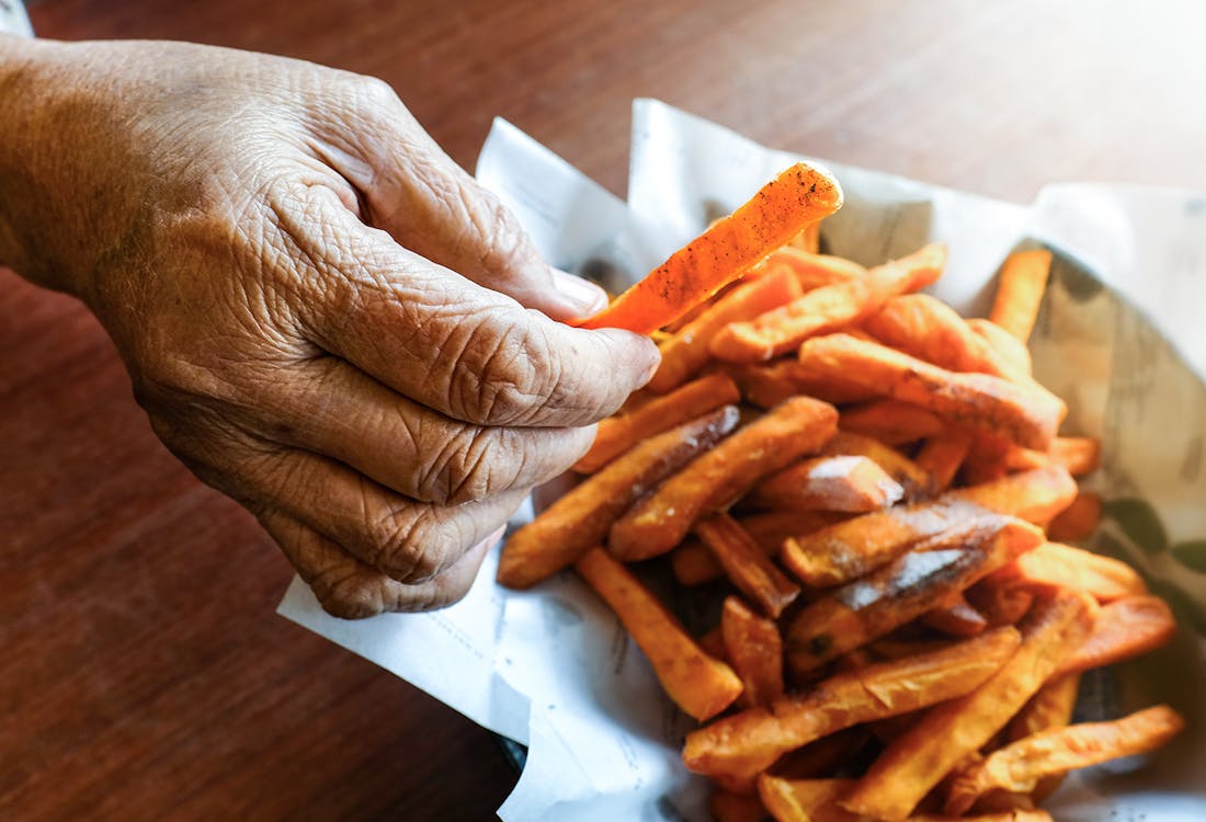 Elderly hand holding crispy sweet potato fry over a bowl of fried sweet potatoes.