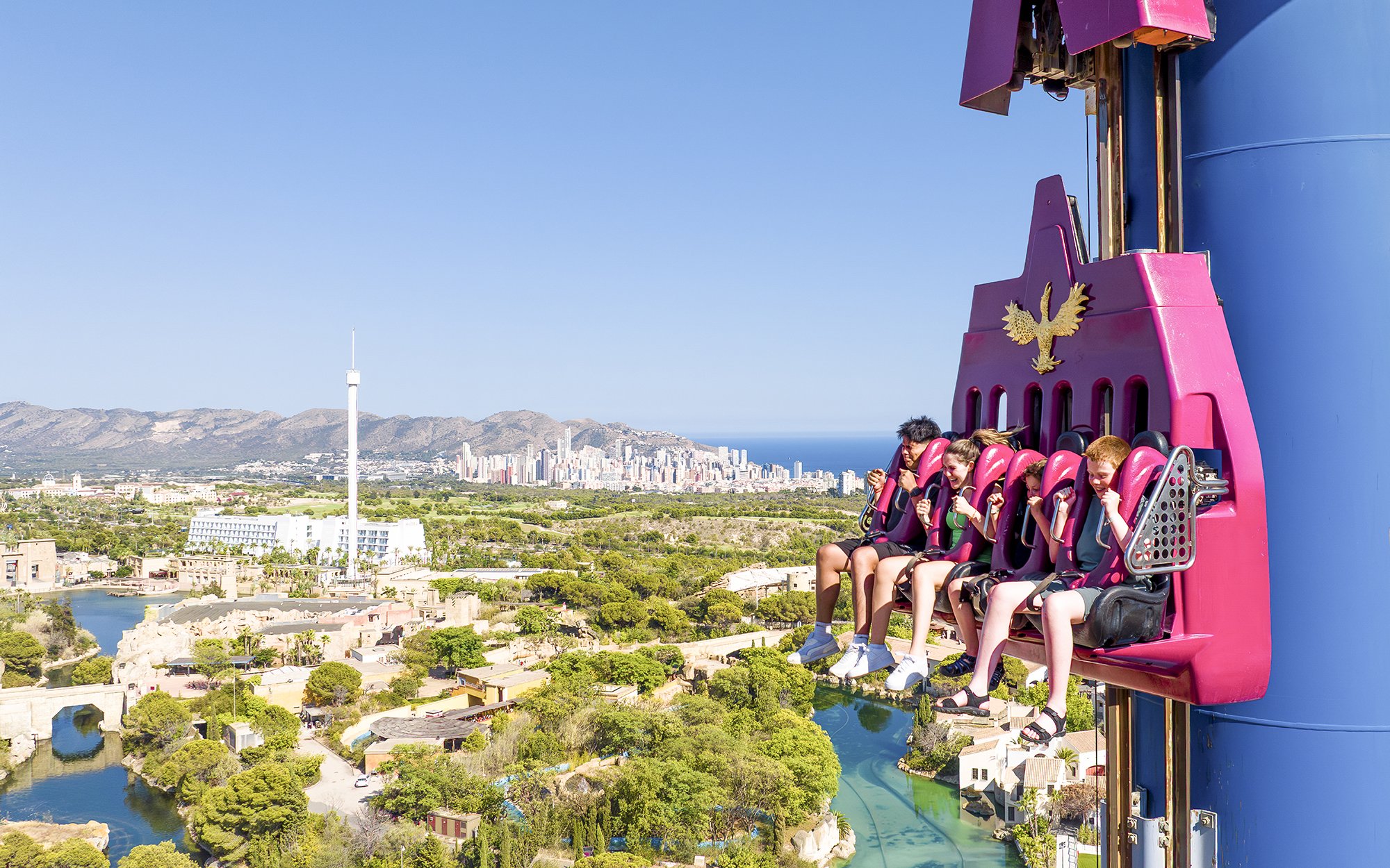 Visitors on a drop tower ride at Terra Mitica Benidorm with cityscape in the background.