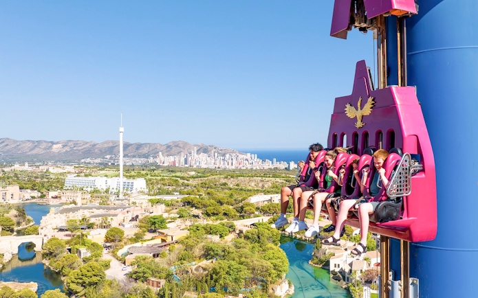 Visitors on a drop tower ride at Terra Mitica Benidorm with cityscape in the background.