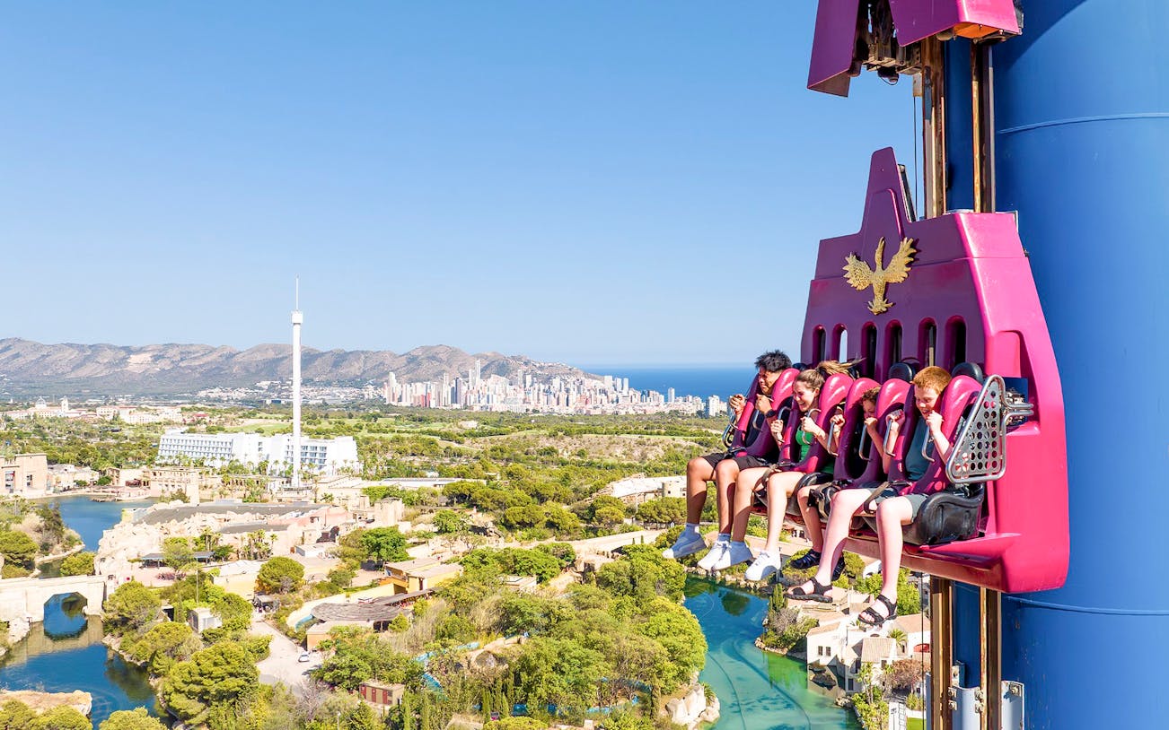 Visitors on a drop tower ride at Terra Mitica Benidorm with cityscape in the background.