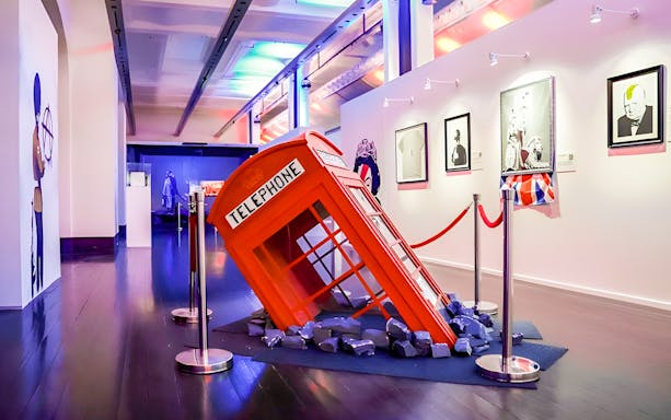Banksy exhibit with a tilted red telephone booth installation and framed artworks.