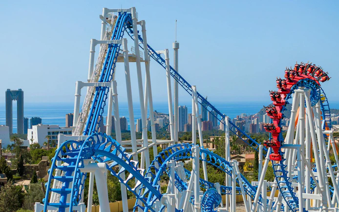 Roller coaster at Terra Mitica theme park in Spain with city skyline in background.