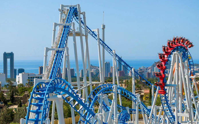 Roller coaster at Terra Mitica theme park in Spain with city skyline in background.