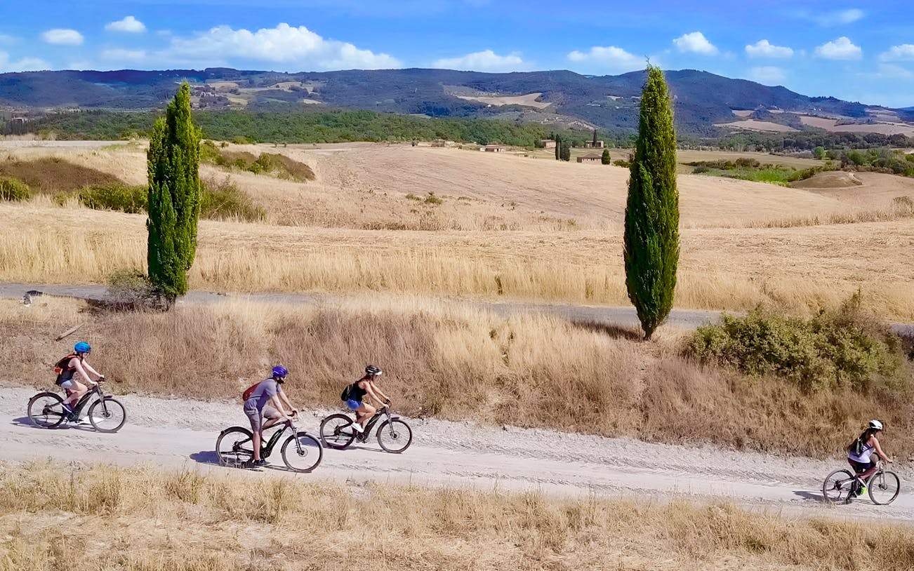 Cyclists on e-bikes ride through Chianti countryside near Florence, flanked by cypress trees.