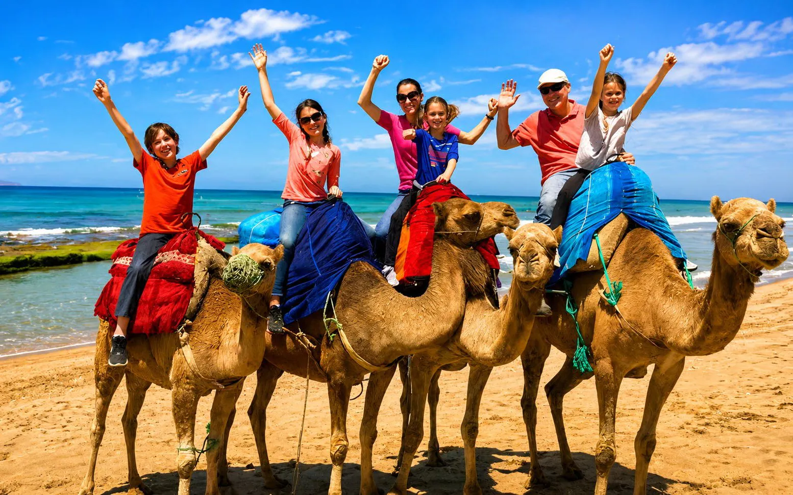 Tourists riding camels on Tangier beach with ocean in background.