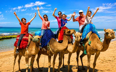 Tourists riding camels on Tangier beach with ocean in background.