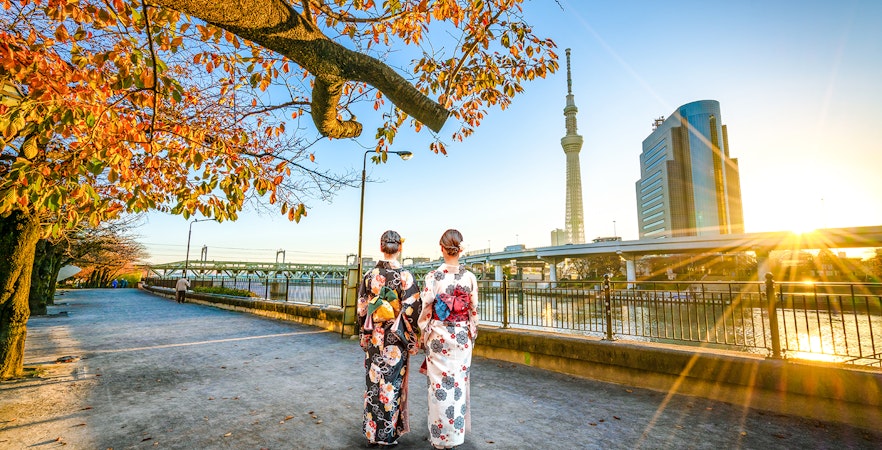Two women in kimonos walking along Sumida River with Tokyo Skytree in the background.