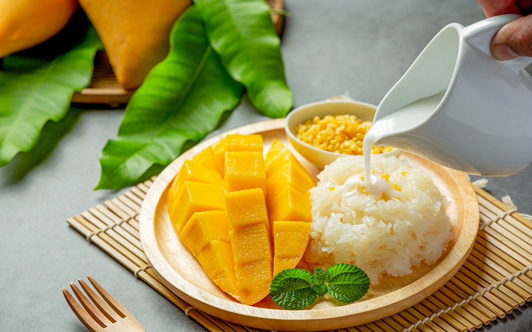 Mango sticky rice with coconut milk being poured, served on a wooden plate.