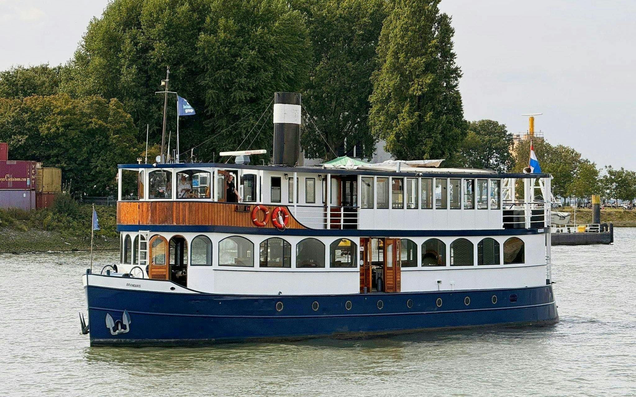 Cruise boat on river for pizza tour with trees and containers in background.
