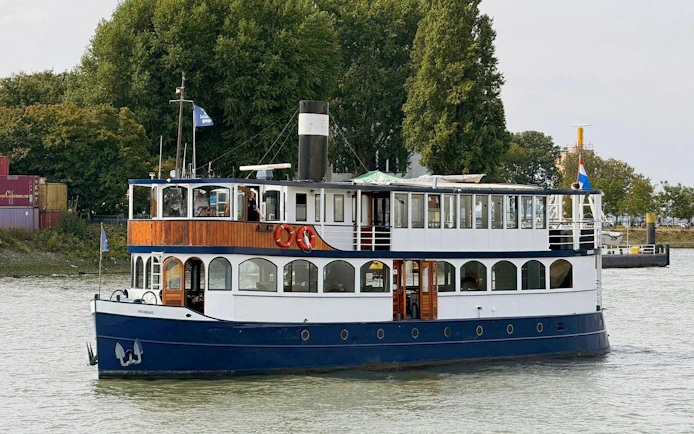 Cruise boat on river for pizza tour with trees and containers in background.