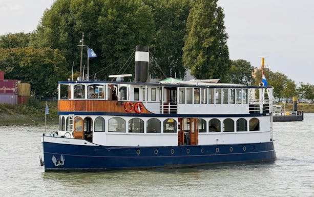 Cruise boat on river for pizza tour with trees and containers in background.