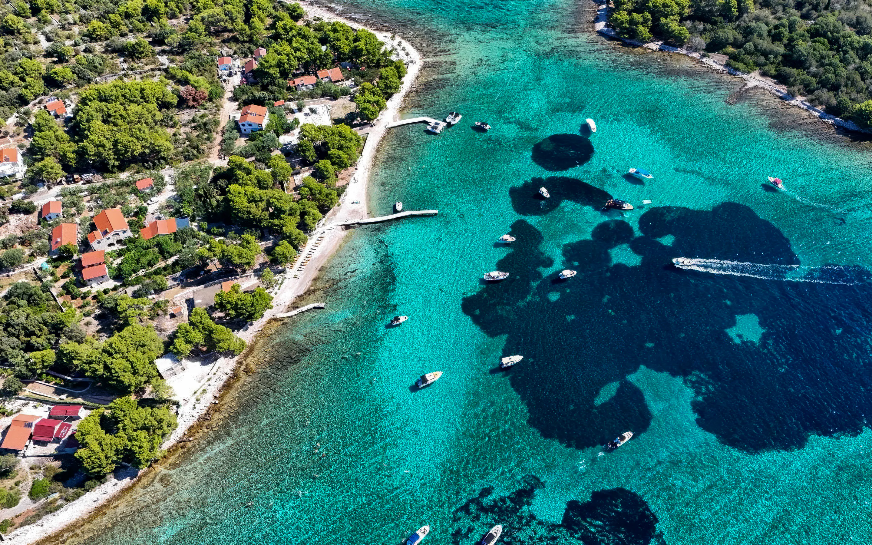 Boats in Blue Lagoon's turquoise waters near Trogir, Croatia, with lush coastline and red-roofed houses.