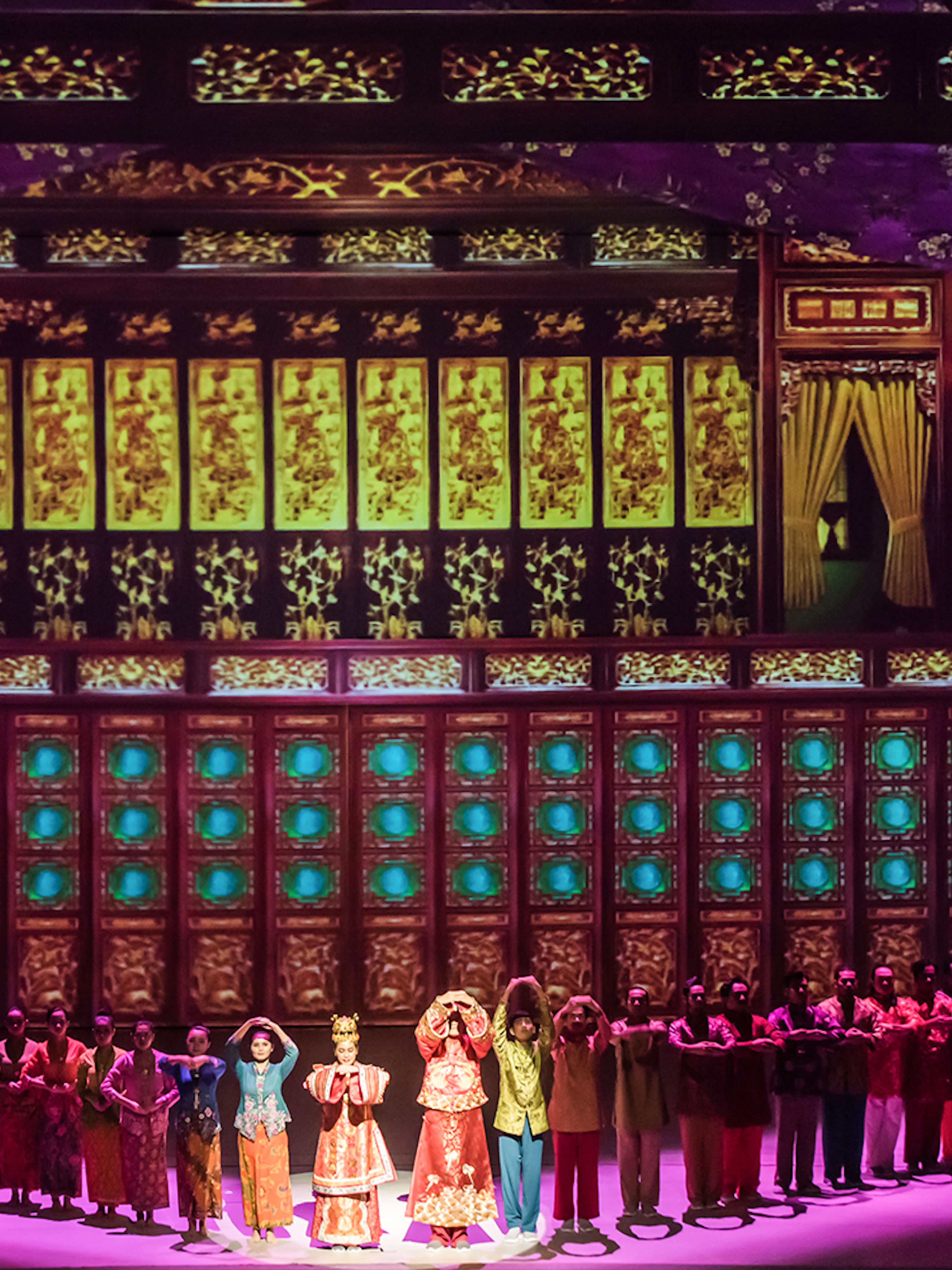 Performers in traditional attire on stage with ornate multi-storey backdrop.