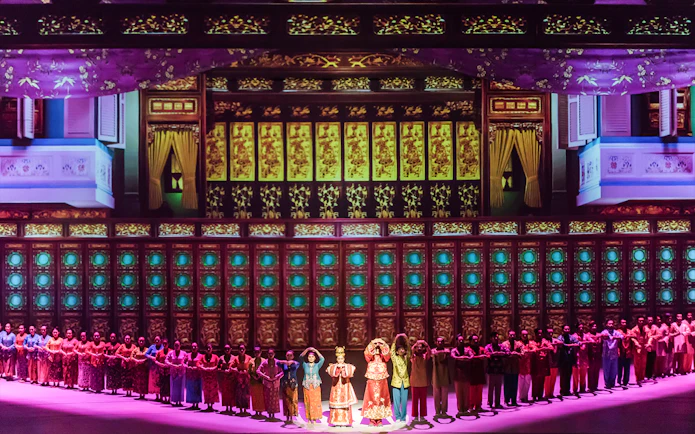 Performers in traditional attire on stage with ornate multi-storey backdrop.