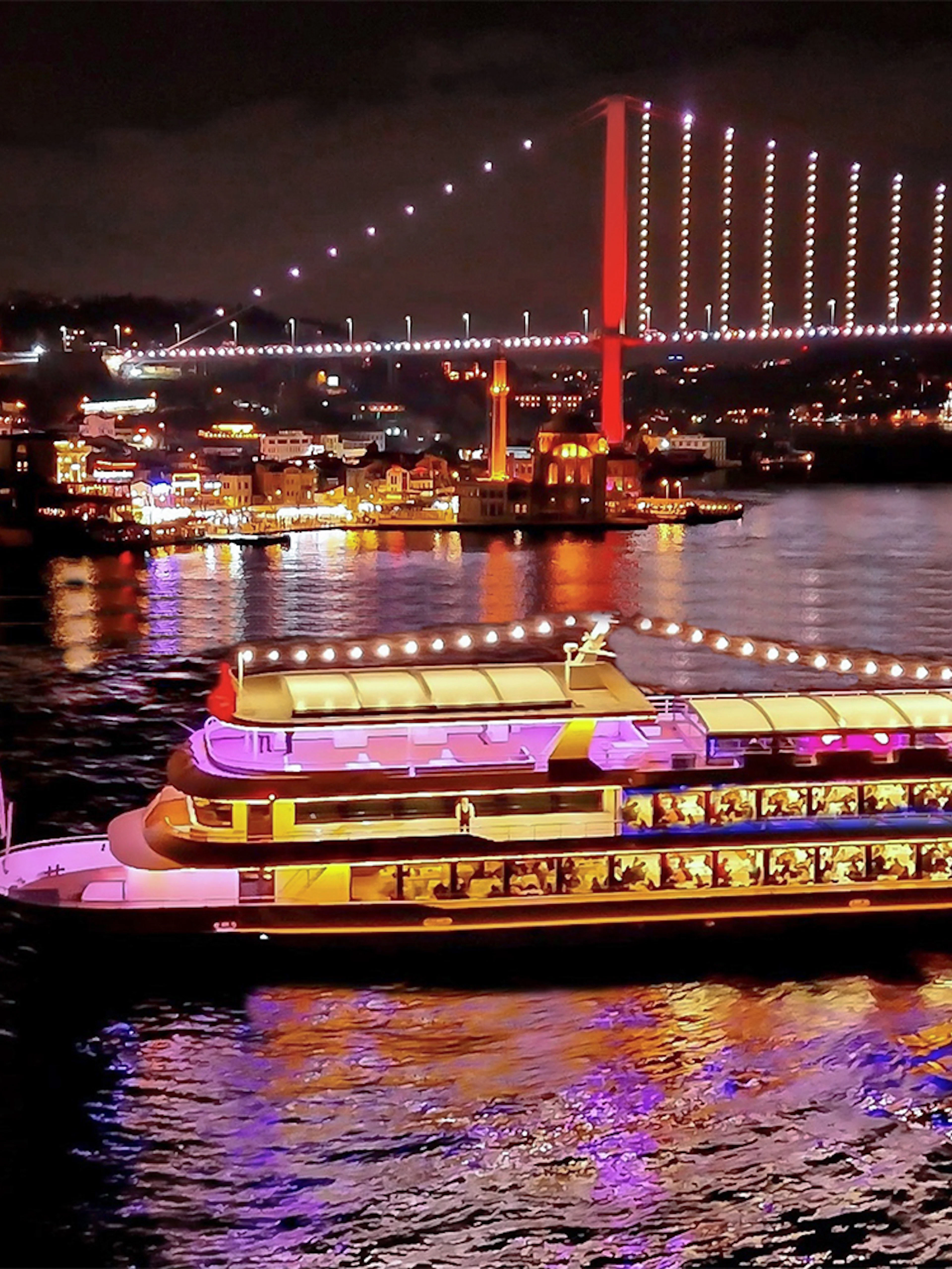 Bosphorus dinner cruise ship illuminated at night with city lights and bridge in Istanbul.