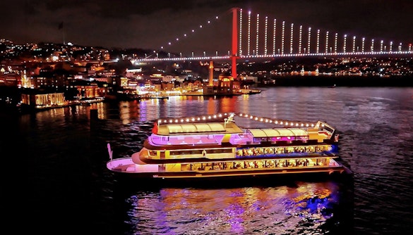 Bosphorus dinner cruise ship illuminated at night with city lights and bridge in Istanbul.