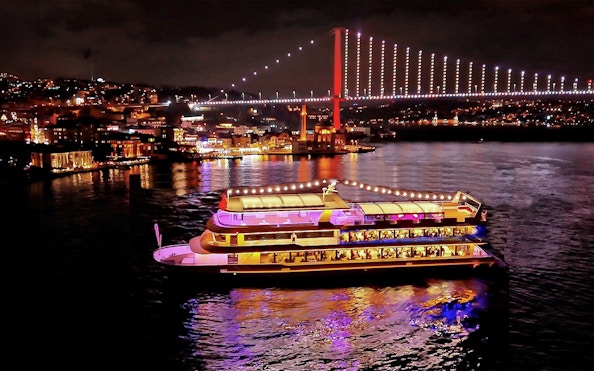 Bosphorus dinner cruise ship illuminated at night with city lights and bridge in Istanbul.