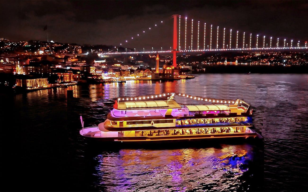 Bosphorus dinner cruise ship illuminated at night with city lights and bridge in Istanbul.