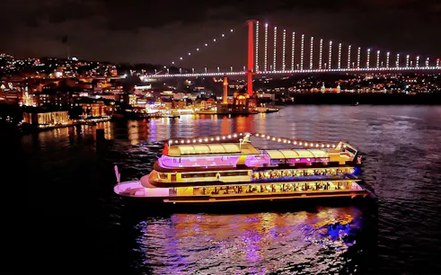 Bosphorus dinner cruise ship illuminated at night with city lights and bridge in Istanbul.