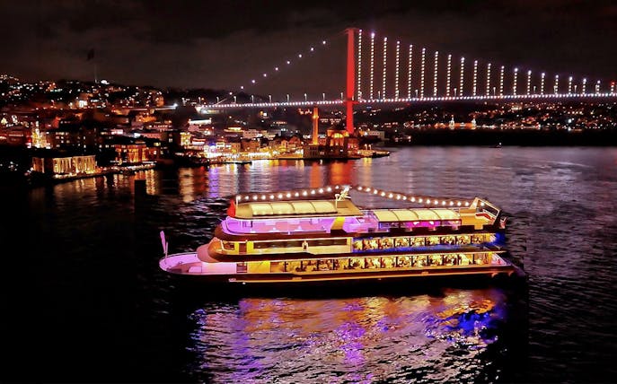 Bosphorus dinner cruise ship illuminated at night with city lights and bridge in Istanbul.