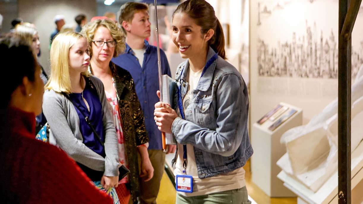 Guide speaking to group near Statue of Liberty, New York City, with Ellis Island in background.